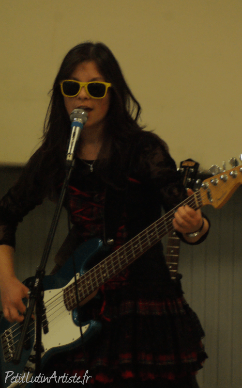 Photo où je chante en jouant de la basse. J'ai des lunettes de soleil jaunes, une robe noire et rouge, et la basse est bleue et possède 5 cordes.
