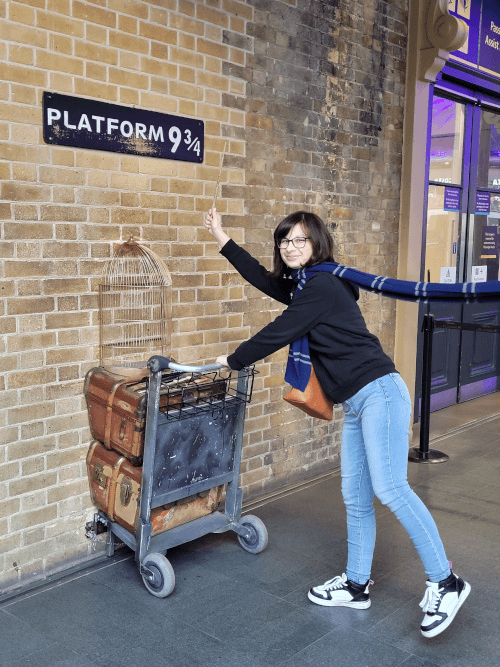 Photo de moi en train de pousser un chariot à valise comme dans Harry Potter, avec une écharpe bleue de Serdaigle, et avec une baguette magique à la main. Je souris en regardant l'objectif.