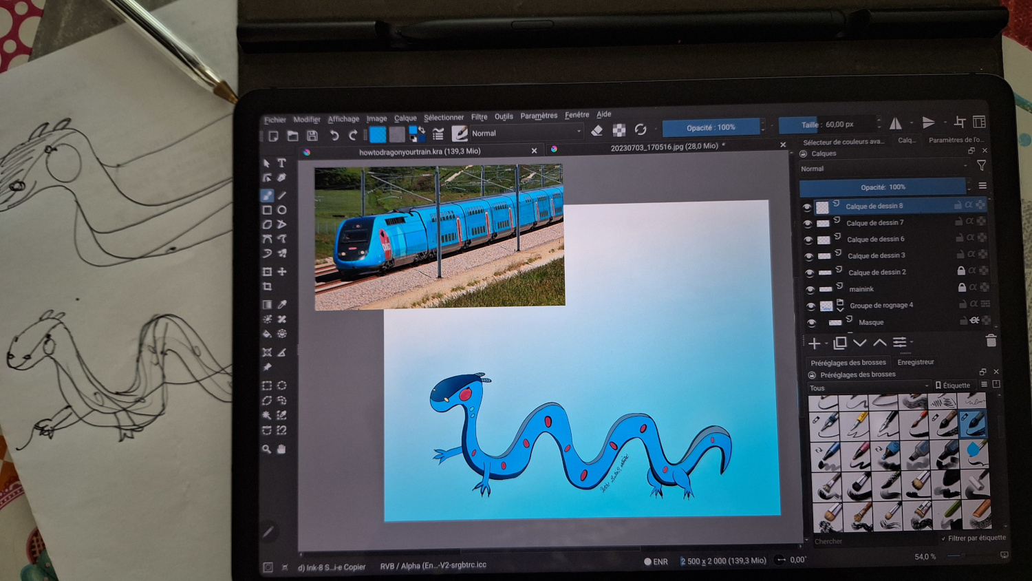 à gauche le croquis sur papier, à droite l'illustration finale sur une tablette samsung galaxy tab s7, faite avec Krita. L'image de référence du train est épinglée en haut à gauche de Krita, et l'illustration finale représente un dragon en longueur allongé avec de petites pattes, il est bleu avec des joues roses.